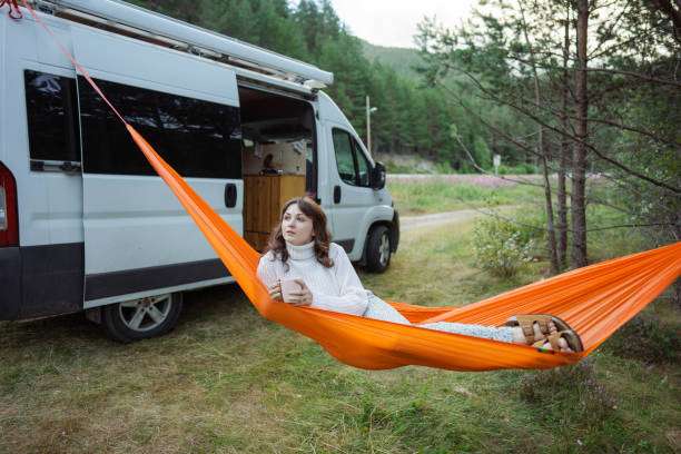 Woman relaxing in a bright orange hammock by a camper van in the forest.