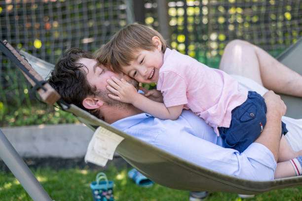 Smiling child hugs father relaxing in a cozy backyard hammock, enjoying family time.