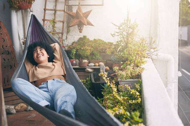 Happy woman relaxing in a grey hammock on a sunny, plant-filled balcony.