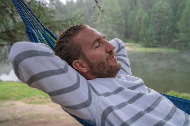 Man with beard peacefully napping in a blue outdoor hammock by a tranquil lake.