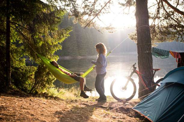 Couple enjoys sunset camping by a lake; one relaxes in a green hammock.