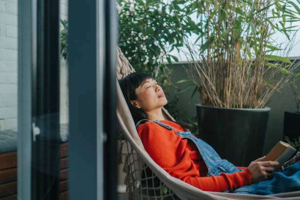 Woman peacefully relaxing, reading a book in a white net hammock on a balcony.