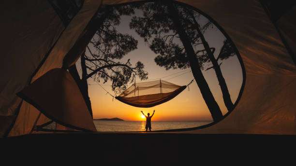 Tent view of a person at sunset near the ocean with an empty hammock between trees.