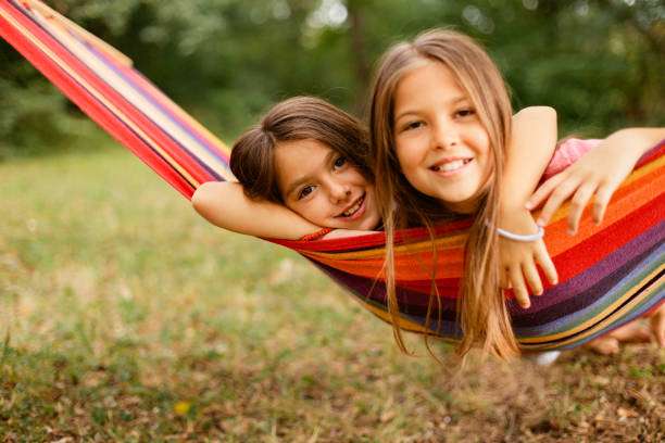Two happy young girls smiling together in a colorful striped hammock outdoors.