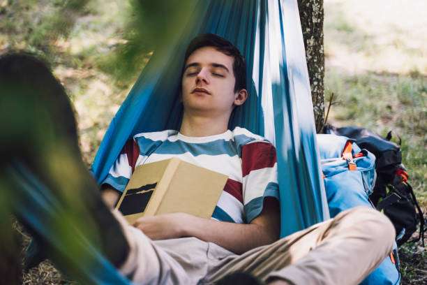 Young man sleeping peacefully in a blue hammock, holding a book, with a backpack nearby.