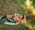 Top view of a Japanese woman enjoys a serene moment outdoors, lounging in a green hammock amidst a tranquil forest setting while holding a mobile phone.