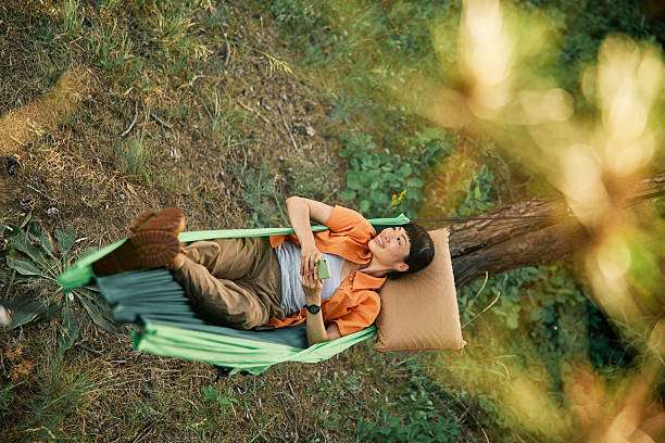 Top view of a Japanese woman enjoys a serene moment outdoors, lounging in a green hammock amidst a tranquil forest setting while holding a mobile phone.